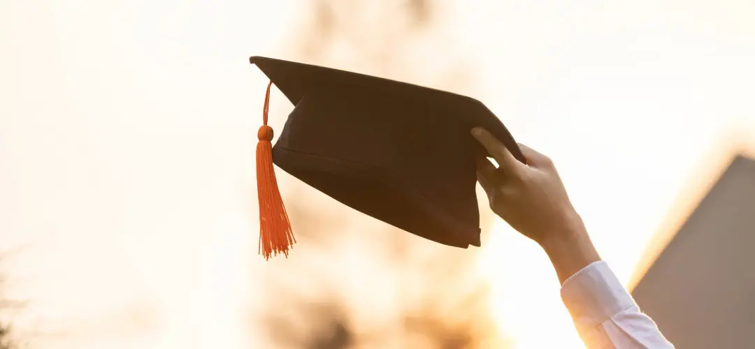 An accelerated bachelor's degree completion student raises their graduation cap in the air against a sunset sky